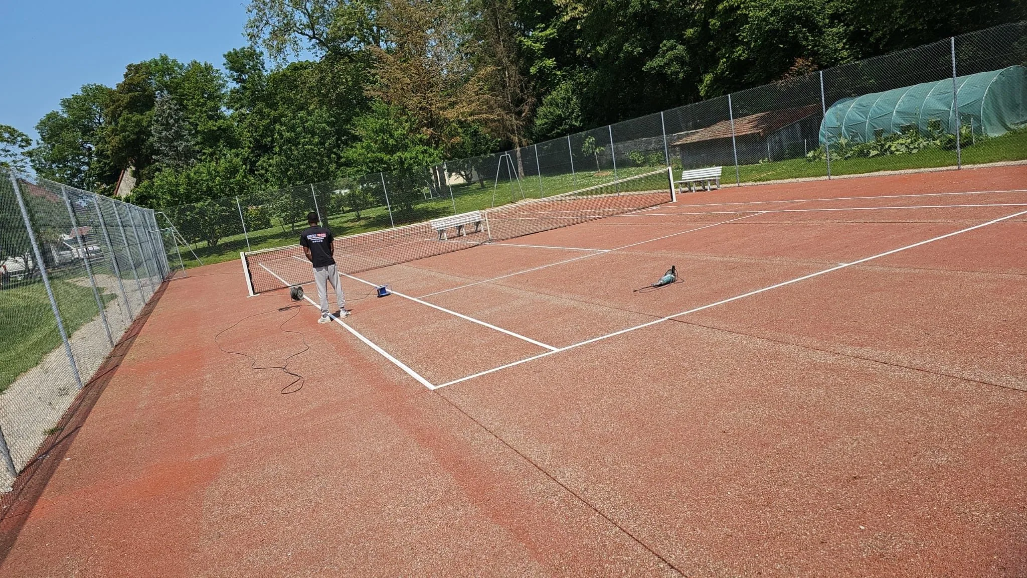 Court de tennis rouge en cours de nettoyage, entouré d'un grillage